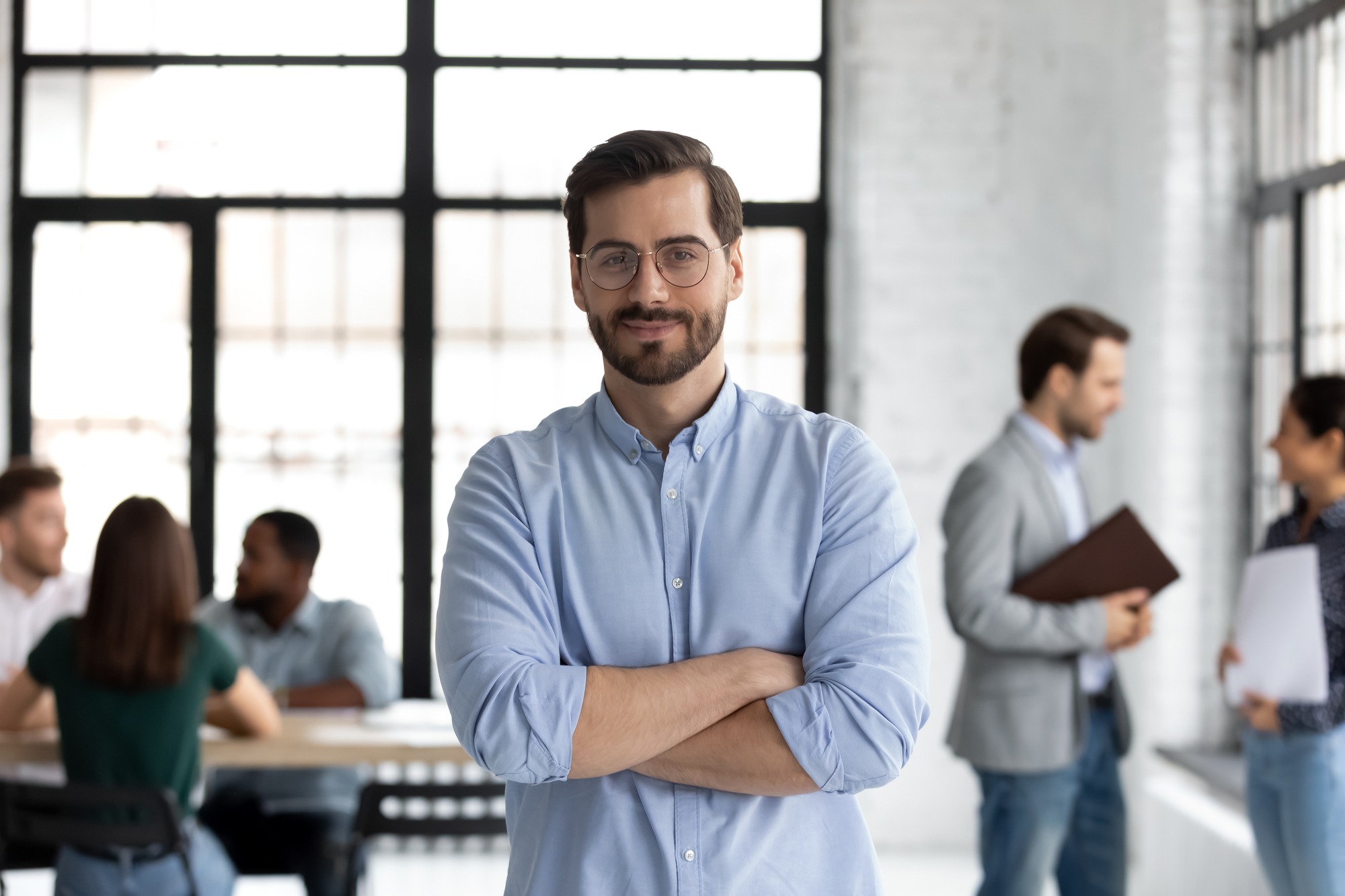 Head shot portrait smiling confident businessman wearing glasses standing in modern office room with arms crossed